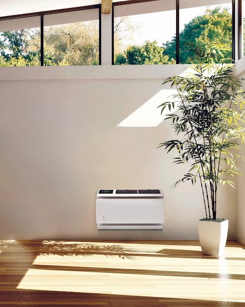 Console-style AC unit installed on a bright wall in a sunlit room with large windows and a tall potted plant