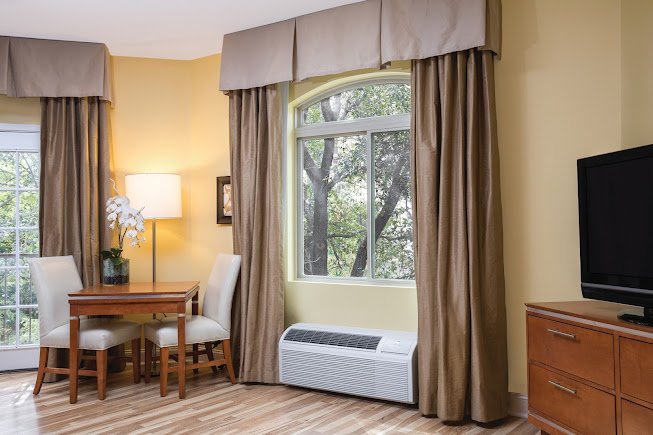 A packaged terminal air conditioner (PTAC) installed below a window in a cozy living area with drapes and seating.